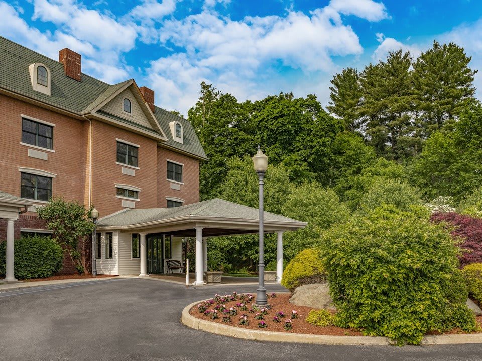 A brick hotel with a covered entrance surrounded by lush greenery and trees under a partly cloudy sky.
