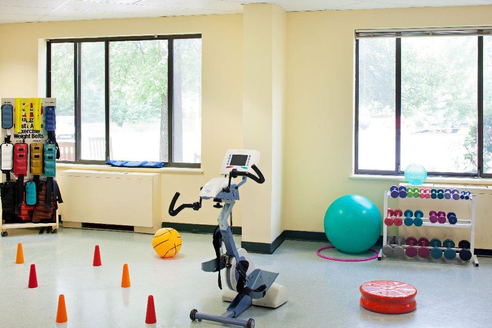 A well-lit exercise room featuring a stationary bike, therapy balls, and training cones.