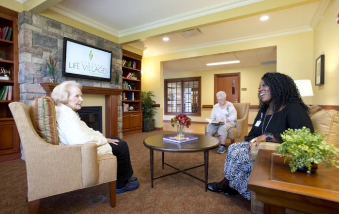 A group of people sitting and conversing in a senior living community room.