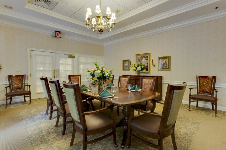 A traditional dining room featuring a wooden table, elegant chairs, and a chandelier overhead. The room is decorated with flowers and paintings on the walls.