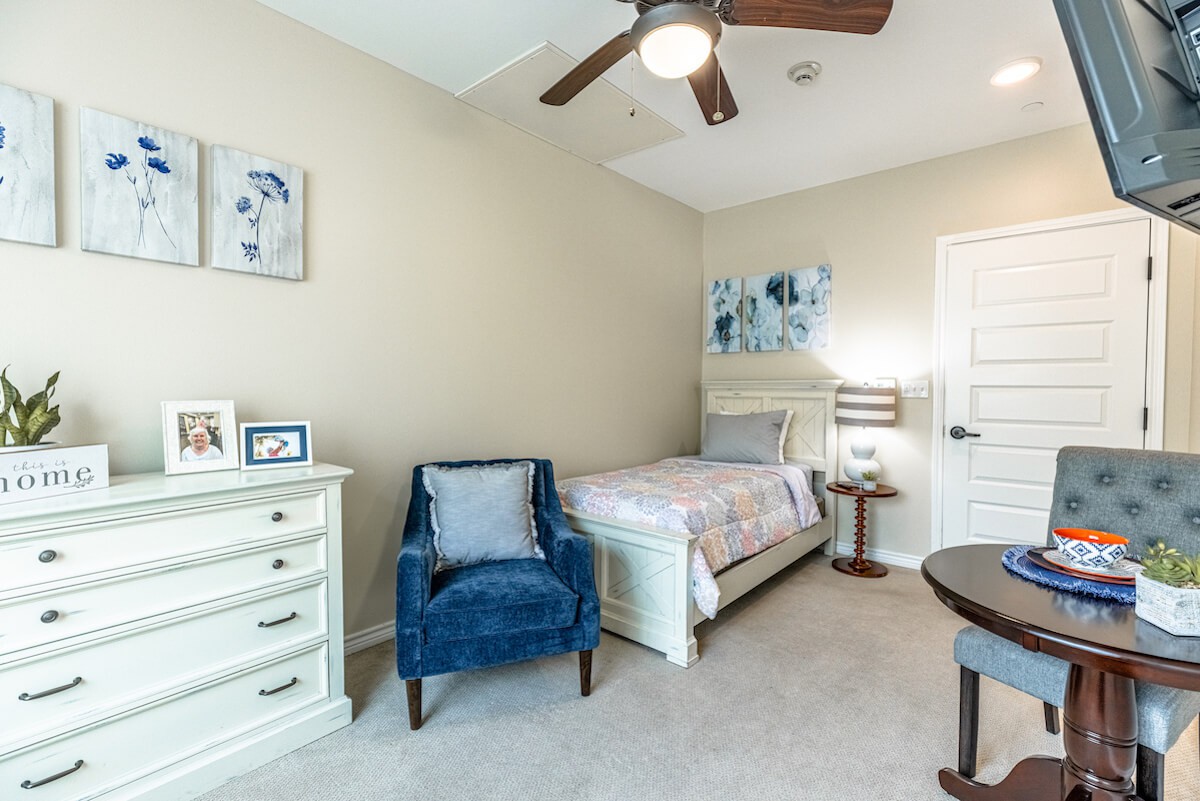 A cozy bedroom with a single bed and a blue armchair, featuring a ceiling fan and decorative artwork.