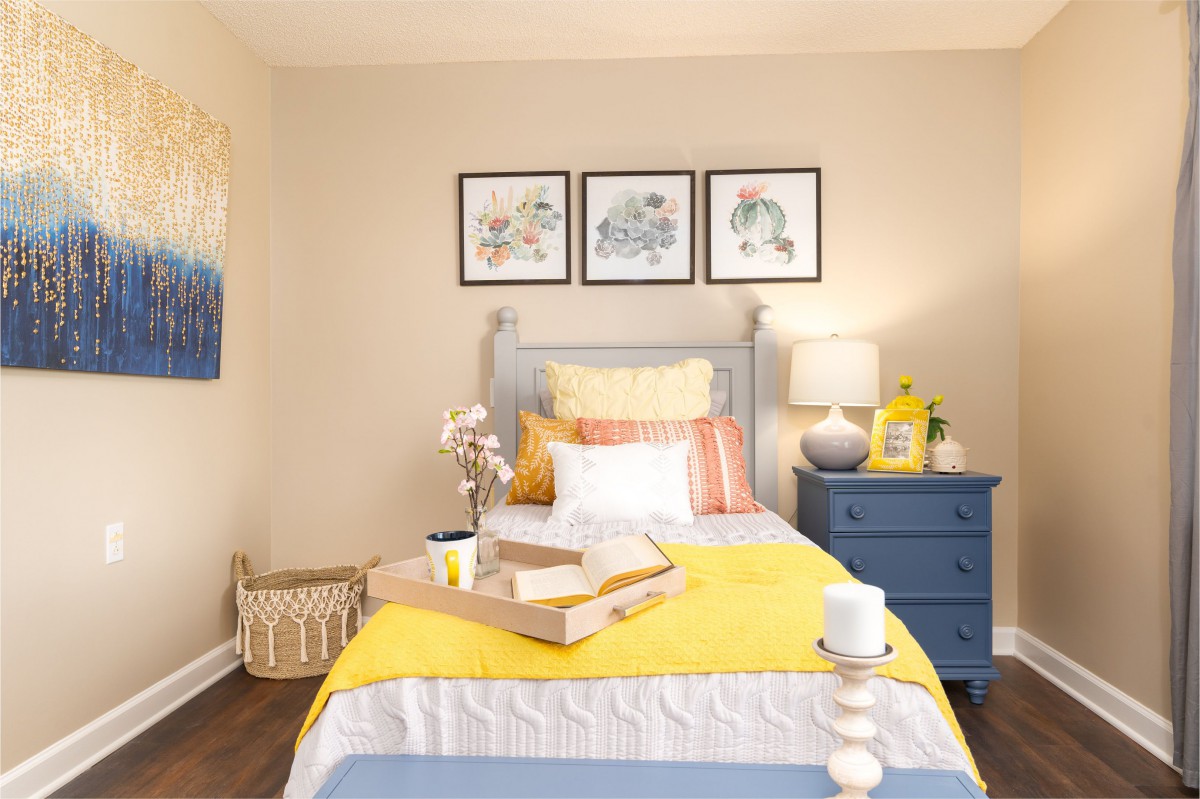 A cozy bedroom with yellow bedding and a blue dresser, complemented by framed wall art and a table lamp.