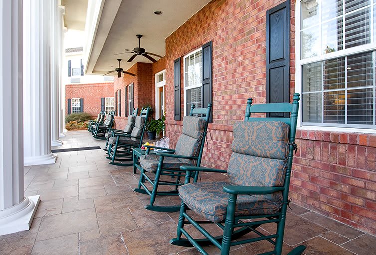 A row of rocking chairs with cushions on a porch against a brick wall, creating a relaxing outdoor seating area.