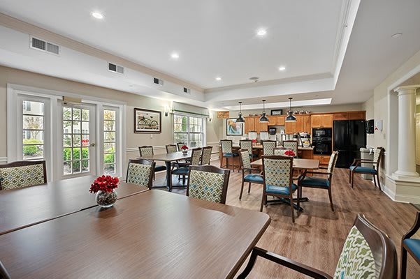 A spacious and well-lit dining area with a kitchen in the background, featuring multiple tables and chairs on a wood floor.