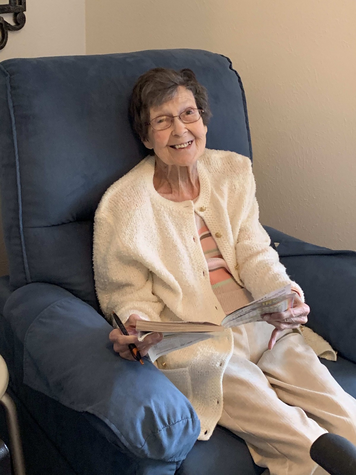 An elderly woman smiling while seated in a chair, holding a book.