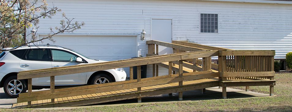 A wooden accessibility ramp leads to the entrance of a house next to a garage with a parked car.