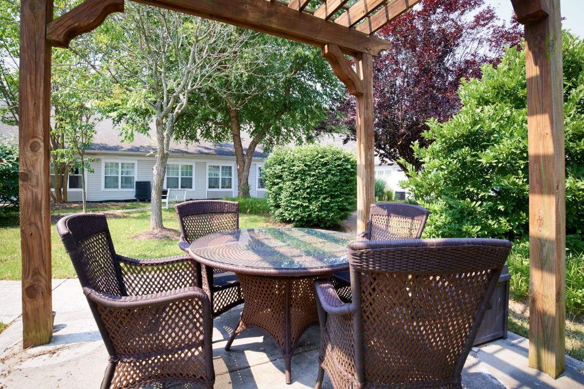 A cozy outdoor patio area featuring a round table with wicker chairs under a pergola, surrounded by lush greenery.