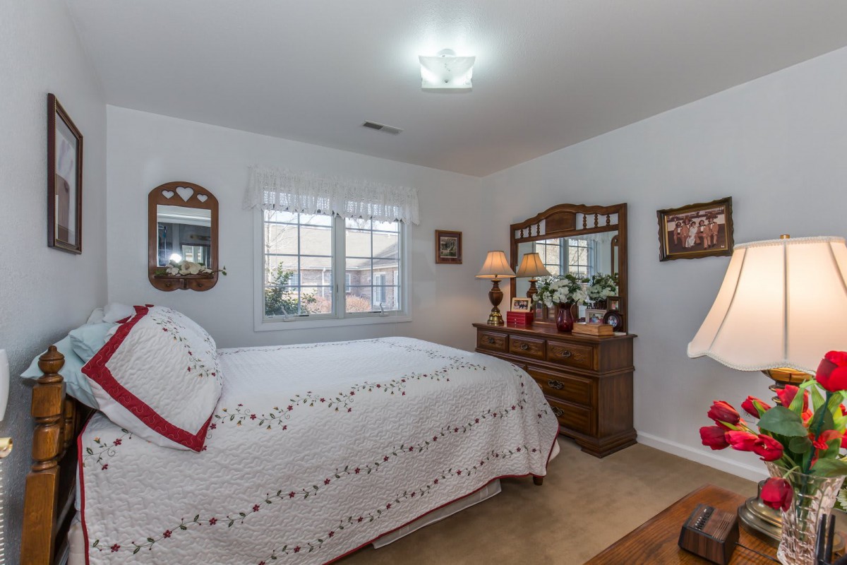 A cozy bedroom with a neatly made bed, wooden dresser, mirrors, and soft lighting.