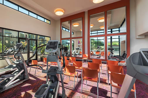 A modern gym with exercise equipment and seating area, featuring large mirrors and windows.