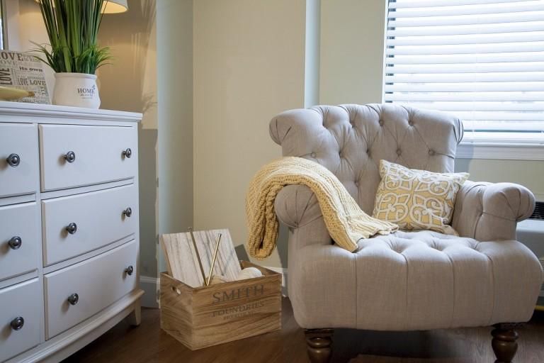 A cozy living room corner with a tufted armchair, soft blanket, and decorative pillow beside a wicker basket and dresser.