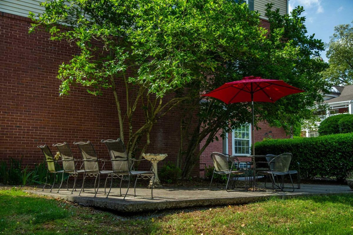 A cozy outdoor patio with seating under a red umbrella and lush green trees.