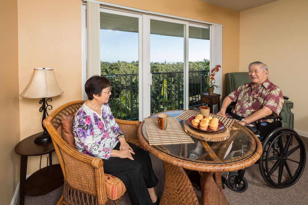 Two seniors relaxing and conversing in a cozy seating area with a scenic balcony view.