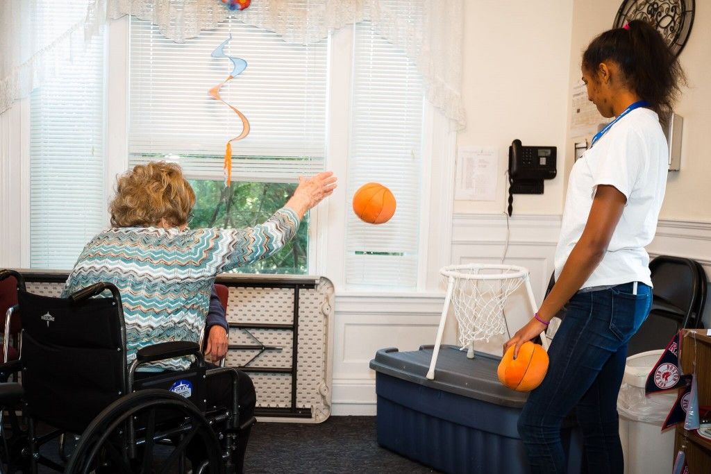 An indoor activity in a care center where an elderly person in a wheelchair participates in a ball toss game with assistance.