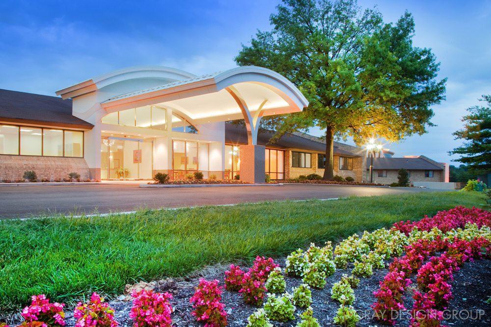 A well-lit building entrance surrounded by colorful flowers and greenery in the evening.