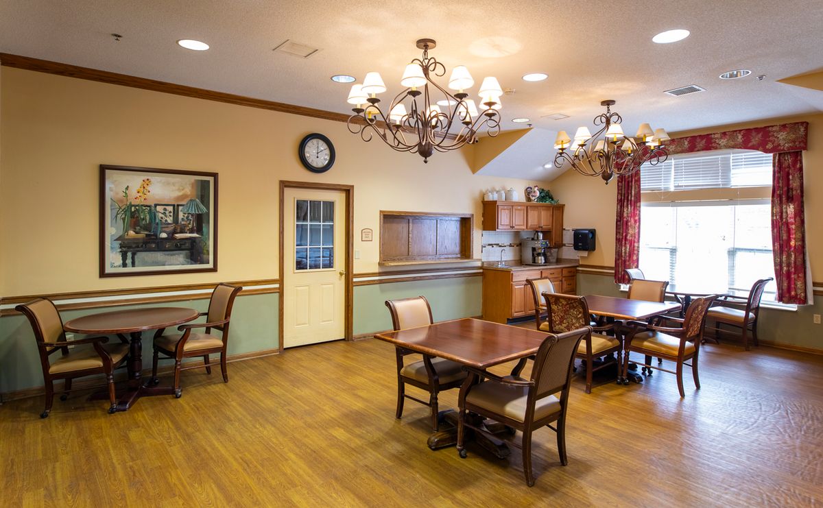 A cozy dining room with wooden flooring and elegant chandeliers, featuring neatly arranged chairs and tables.