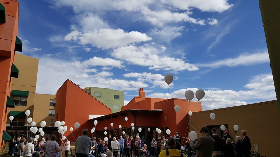 A lively outdoor event with people holding balloons under a bright blue sky in front of uniquely shaped buildings.