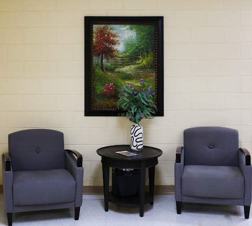 A cozy waiting area featuring two gray chairs, a small table with a decorative vase, and a nature-themed painting on the wall.