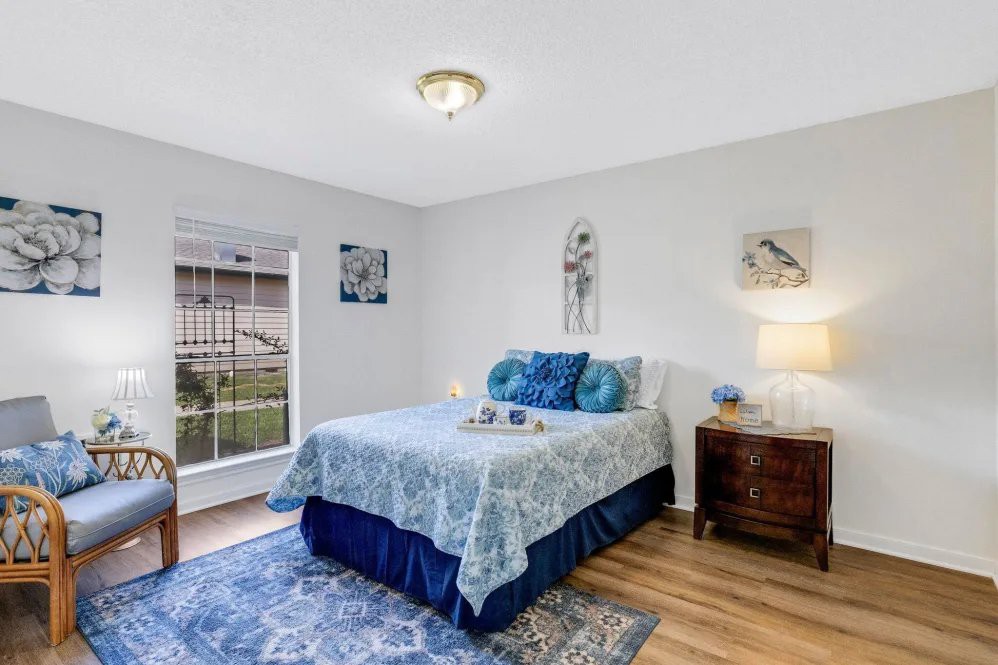 A cozy bedroom with a double bed, featuring blue and white bedding, surrounded by wall art and a nightstand with a lamp.
