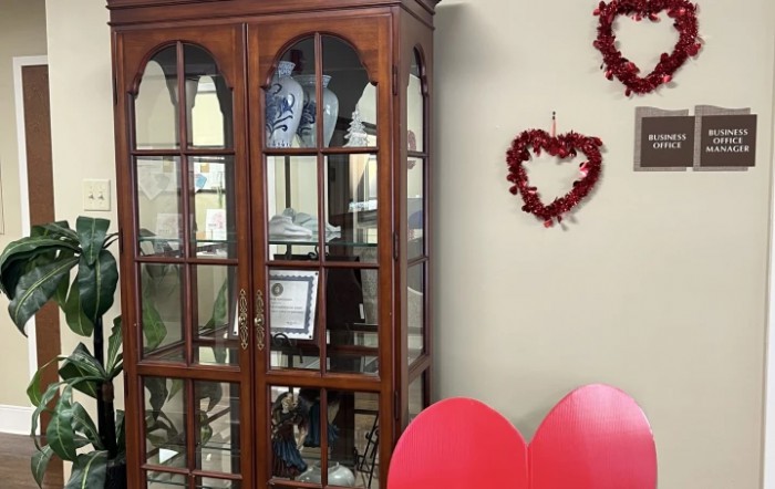 A room interior featuring a wooden cabinet with glass doors, housing various vases and decorative items. Red heart decorations adorn the adjacent wall.