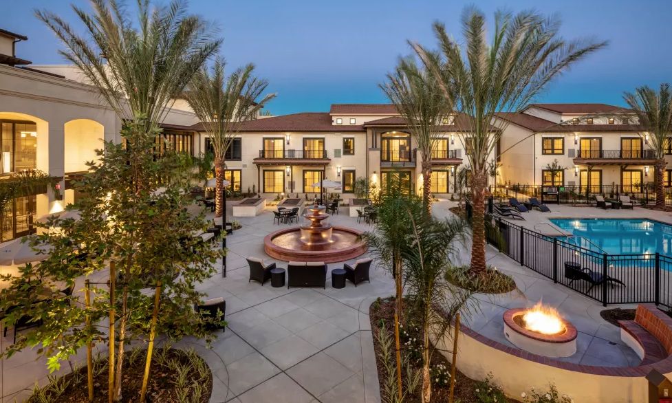 A luxurious residential building with a courtyard featuring a fountain, surrounded by palm trees and outdoor seating in an evening setting.