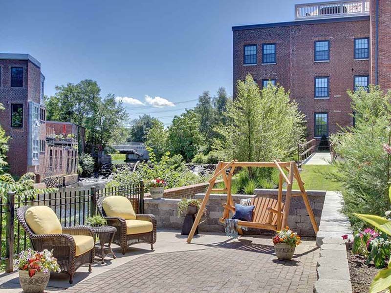 A sunny outdoor patio with comfortable seating and a wooden swing, set against a backdrop of greenery and a brick building.