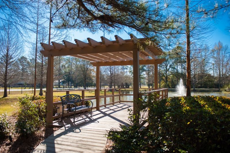 A serene park scene featuring a wooden pergola and bench overlooking a pond with a fountain.