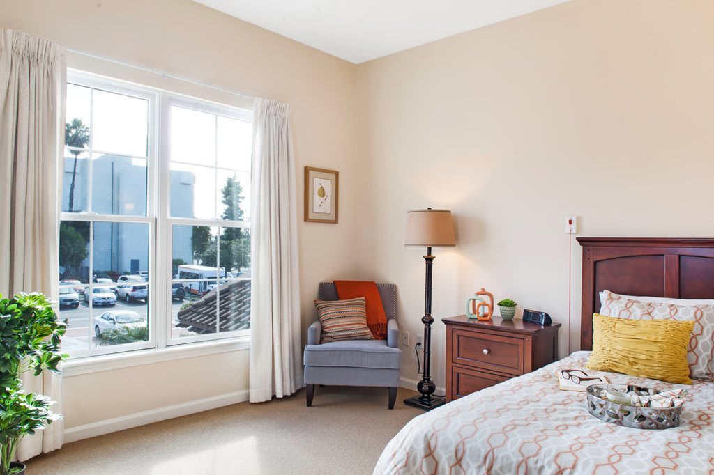 A cozy bedroom featuring a comfortable chair by the window and a bedside lamp.