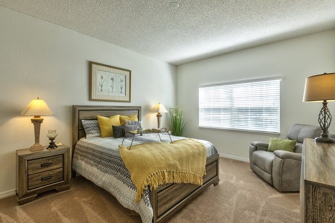 A cozy and well-decorated bedroom featuring a bed with yellow and gray accents, a lamp on a nightstand, and a soft carpet underfoot.