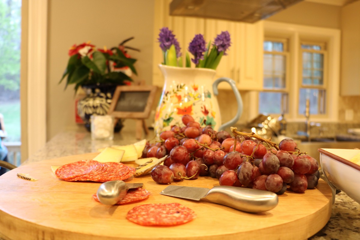 A kitchen scene with grapes, salami, and cheese on a cutting board, accompanied by a decorative pitcher with flowers.