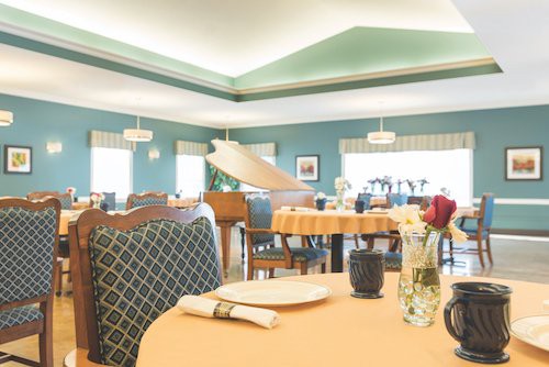 A bright dining room with neatly arranged tables, chairs, and a decorative piano.