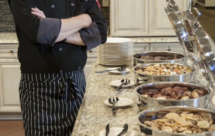 A chef standing beside a buffet setup with various dishes and plates in a kitchen setting.