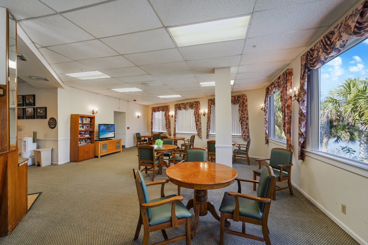 A cozy living area featuring round tables, chairs, a television, and large windows with drapes.
