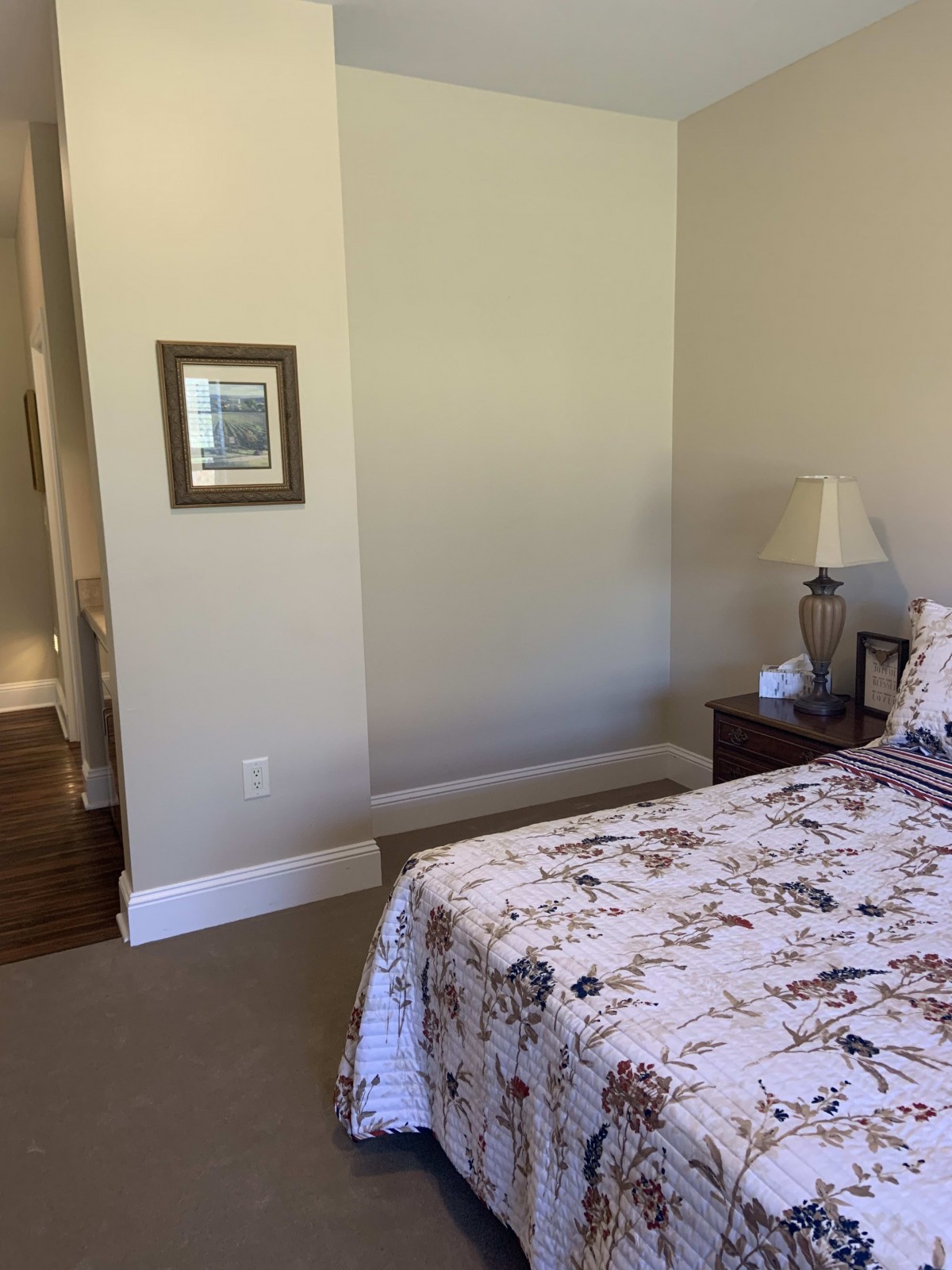 A cozy bedroom with a floral bedspread, a nightstand with a lamp and framed photograph, and a framed picture on the wall.