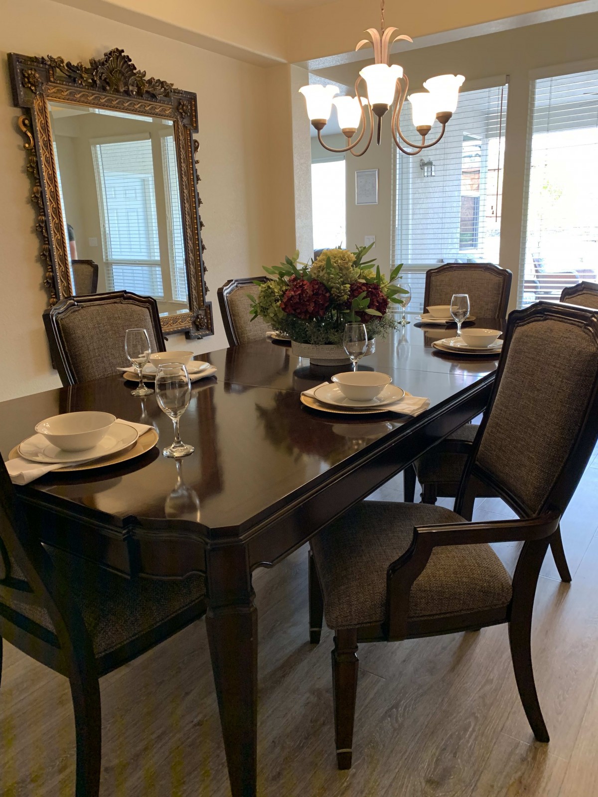 A stylish dining room featuring a dark wood table set with elegant chairs, complemented by a large mirror and chandelier.