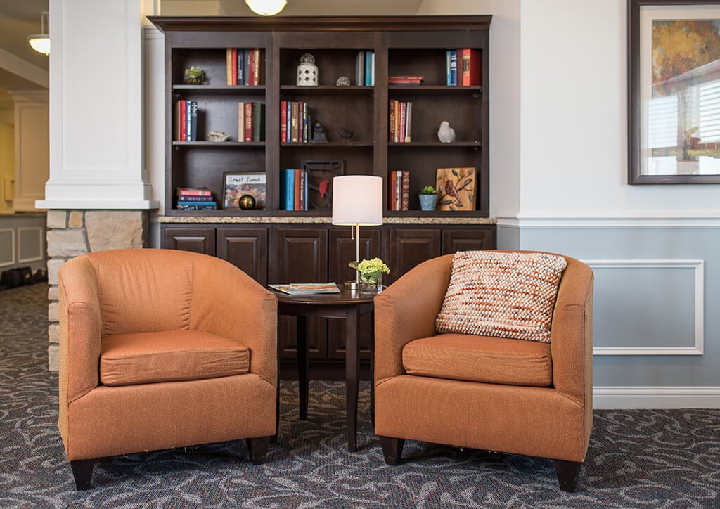 A cozy living room space featuring two brown armchairs, a wooden bookshelf filled with books and decor, and a small side table with a lamp.