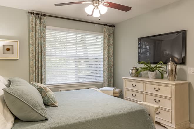 A cozy bedroom with a large window, neatly made bed, and a dresser with decorative items.