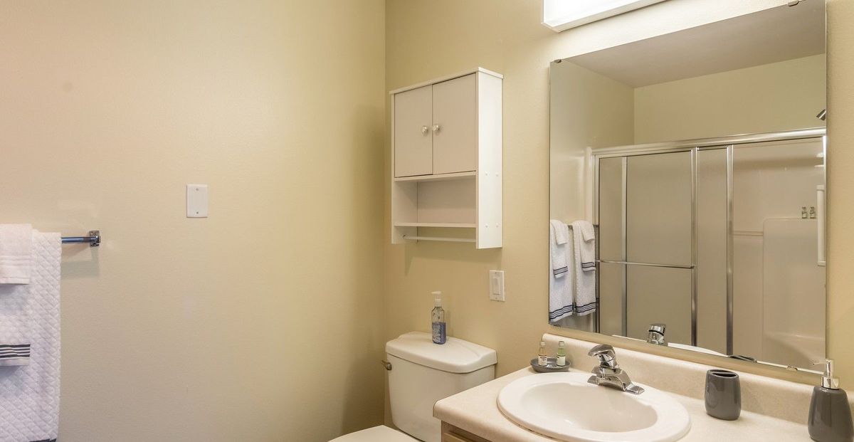 A small bathroom featuring a sink with a mirror, cabinet, and a toilet with towels in the background.
