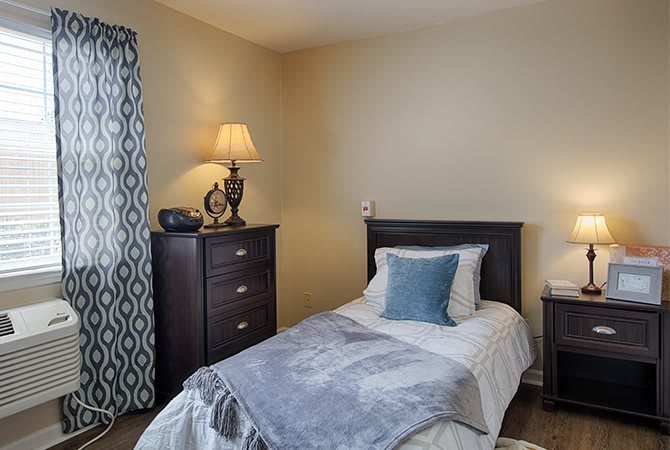 A cozy bedroom with a single bed, adorned with soft pillows and a gray blanket. A lamp sits on a dark wooden dresser, adjacent to patterned curtains.