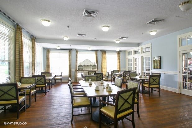 A spacious dining room with multiple tables and chairs, featuring large windows and wooden flooring.