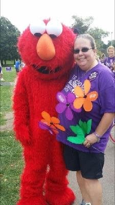 A person in a purple shirt is standing outside next to someone dressed in an Elmo costume, holding colorful flowers.