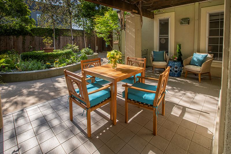 A cozy outdoor patio with a wooden table and chairs, surrounded by lush greenery.