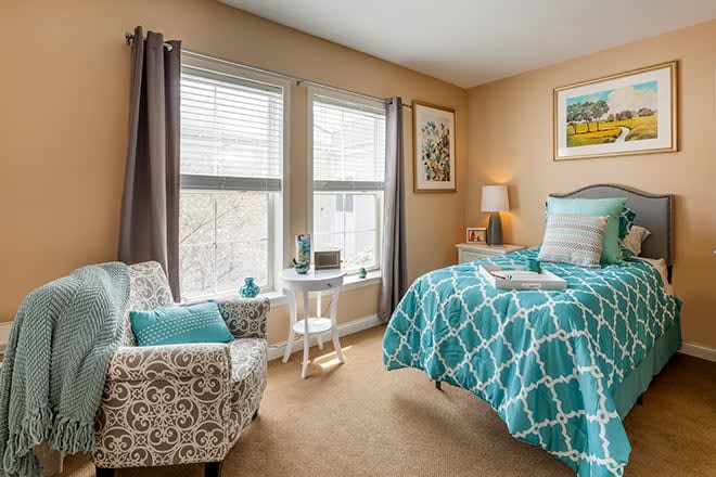 A cozy bedroom with a teal and white patterned bedspread, featuring an armchair, natural light from two large windows, and decorative artwork on the walls.