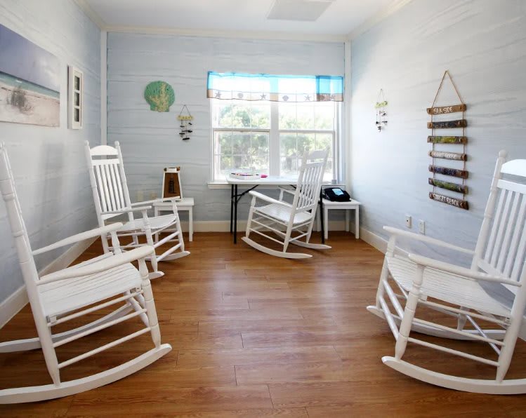 A cozy room with white rocking chairs and a wooden floor, featuring a window and wall decorations.