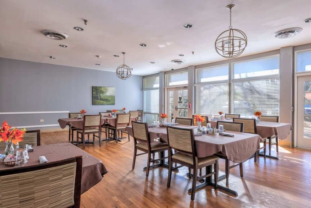 A modern restaurant dining area with neatly arranged tables and chairs, illuminated by natural light through large windows.