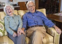 An elderly couple sitting on a couch, laughing and holding hands.