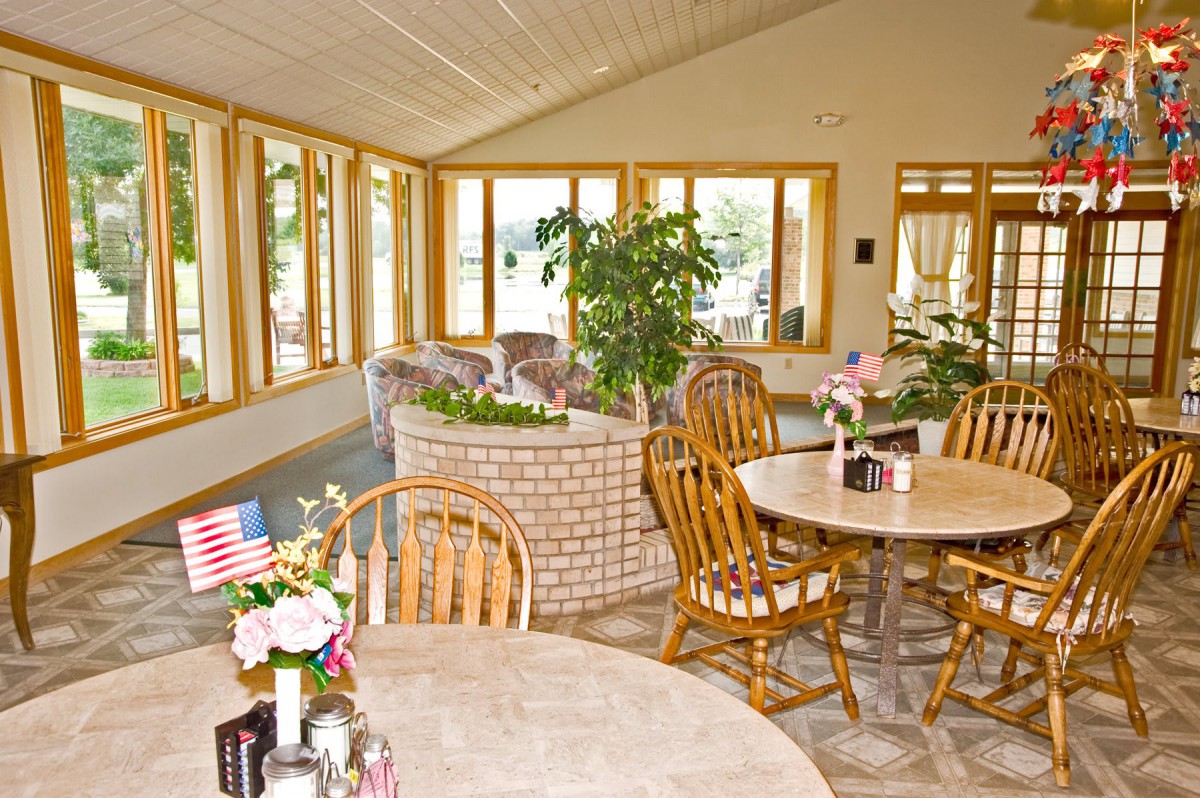 A cozy dining room with wooden tables and chairs, surrounded by large windows and indoor plants.