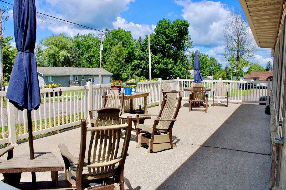 A sunny patio with wooden chairs and tables, surrounded by a white fence and trees in the background.