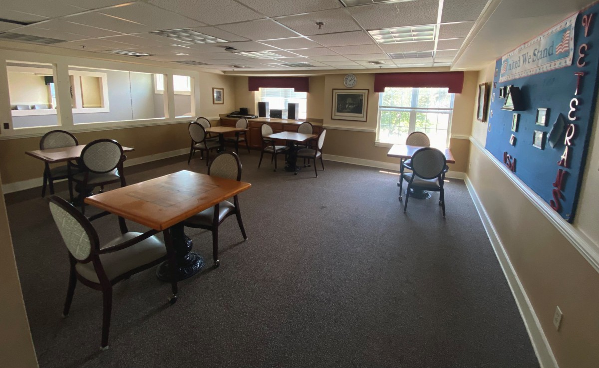 A spacious room with several tables and chairs, featuring large windows and carpeted floors.