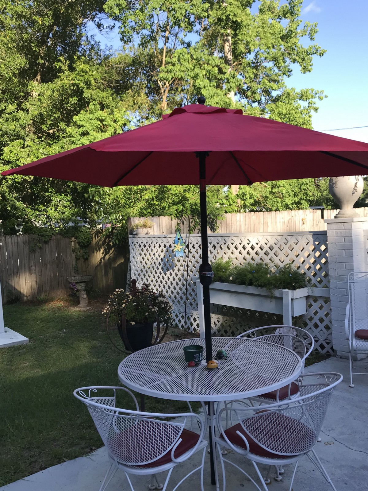 A patio setting with a red umbrella over a round table and chairs, situated in a garden.
