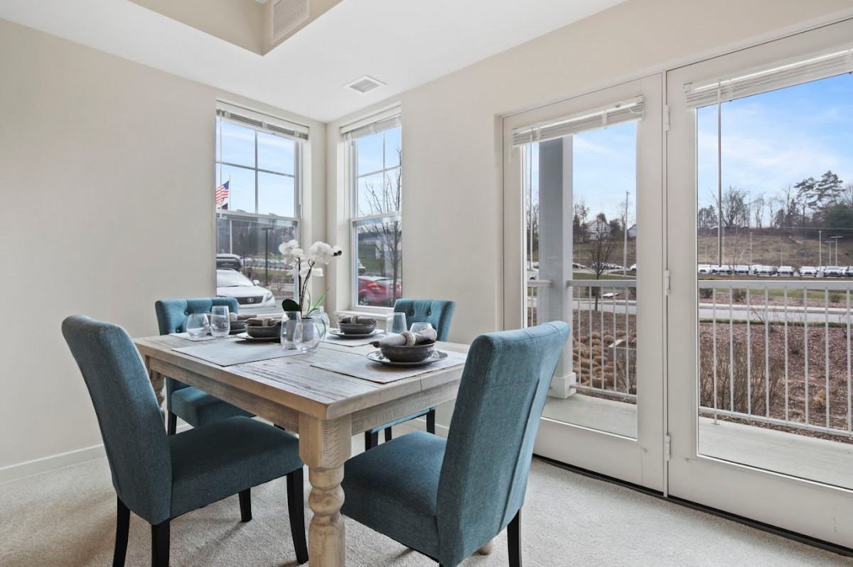 A bright dining area with a wooden table and upholstered chairs, featuring large windows that allow natural light to fill the space.